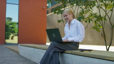 Beyaz gömlekli ve kulaklıklı genç bir kadın video görüşmesi sırasında dışarıda dizüstü bilgisayarıyla otururken konuşuyor..
