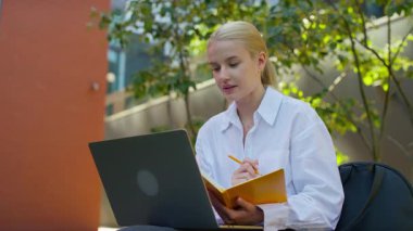 Beyaz tişörtlü genç bir kadın dışarıda online bir derse katılırken dizüstü bilgisayar izliyor ve sarı bir not defterine yazıyor..