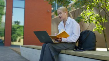 Kulaklık takan genç bir kadın, sarı bir not defterine yazarken ve dışarıda dizüstü bilgisayar kullanırken bir video görüşmesinde konuşuyor..