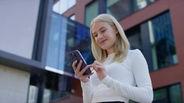 Güneşli bir günde modern cam ve tuğla binaların önünde dikilirken beyaz bluzlu gülümseyen sarışın kadın akıllı telefon kullanıyor..