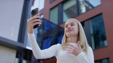 Beyaz bluzlu sarışın kadın akıllı telefon tutuyor ve modern ofis binalarının önünde görüntülü konuşuyor..