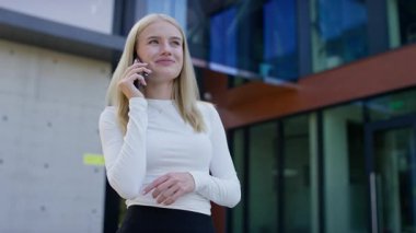 Güneşli bir günde modern bir ofis binasının önünde beyaz bluzlu, kendine güvenen sarışın kadın akıllı telefonuyla konuşuyor..