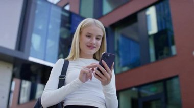 Sırt çantalı, şaşkın ve mutlu genç bir kadın modern binaların önünde telefonuna bakarken gülümseyerek tepki veriyor..