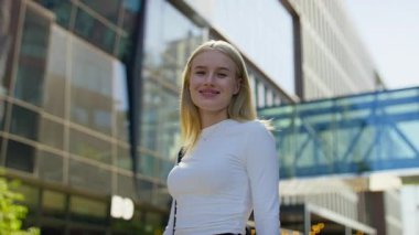 Beyaz bluzlu genç bir kadının portresi günışığında modern ofis binalarının dışındaki kameraya bakarken gülümsüyor..