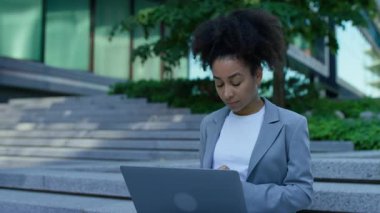 Gri ceketli ciddi bir genç kadın şehir merdivenlerinde finansman ya da bütçe yönetirken dizüstü bilgisayarıyla çalışıyor..