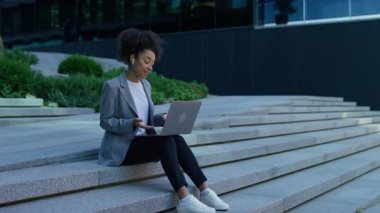 Spor ceketli ve kulaklıklı profesyonel bir kadın, şehir merdivenlerinde oturmuş, dizüstü bilgisayarında bir video görüşmesi yaparken el kol hareketi yapıyor..