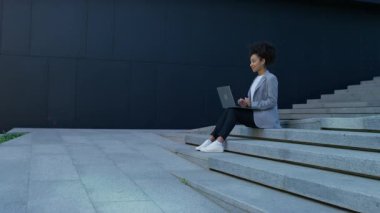 Kendine güvenen, spor ceketli bir kadın şehir basamaklarında oturmuş uzaktan çalışan modern bir girişimciyi temsil ediyor..