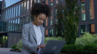 Modern şehir ortamında bir bankta oturmuş gülümseyen genç bir kadın, dizüstü bilgisayar kullanıyor, muhtemelen internette geziniyor ya da alışveriş yapıyor. Dijital bir aktivite sırasında rahat ve memnun görünüyor..