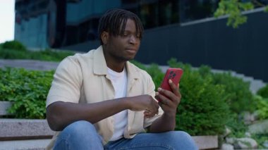 Şehir parkındaki basamaklarda oturan genç adam, akıllı telefonunu kaydırmaya odaklanmış, muhtemelen sosyal medyayı karıştırıyor veya internetten öğrenim görüyor..