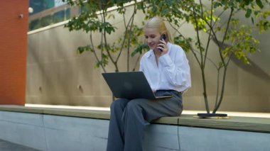 Dışarıda bankta oturan, akıllı telefonuyla konuşan, dizüstü bilgisayarda çalışan, uzaktan kumanda sırasında birden fazla iş yapan profesyonel bir kadın..