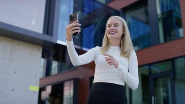 Neşeli genç bir kadın güneşli bir günde modern bir ofis binasının önünde dikilirken akıllı telefonuyla bir video görüşmesi yapıyor..