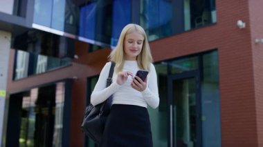 Sırt çantalı genç bir kadın cam ofis binasının önünde duruyor, akıllı telefonunu internette gezinmek ya da çalışmak için kullanıyor..