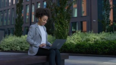 Neşeli genç bir profesyonel modern bir ofis binasının önündeki bankta oturuyor, internette dizüstü bilgisayarla geziniyor ve gülümsüyor..