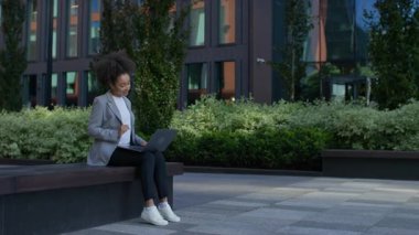 Profesyonel bir kadın modern bir şehir ortamında bankta oturuyor ve dizüstü bilgisayarındaki iş ilanlarını faiziyle karıştırıyor..