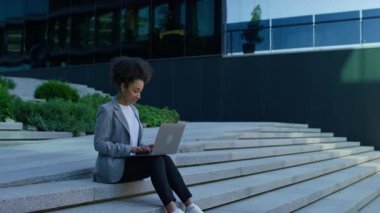 Odaklanmış bir öğrenci binanın dışındaki modern beton basamaklara oturur, sakin, yeşil kentsel bir ortamda dizüstü bilgisayarla çalışır..