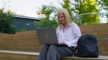 Genç kız öğrenci dışarıda tahta basamaklarda oturuyor, yanında bir sırt çantasıyla dizüstü bilgisayarda geziniyor..
