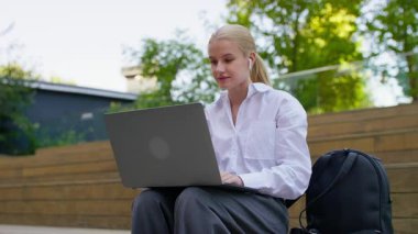 Dıştaki merdivenlerde dizüstü bilgisayarla oturan, kablosuz kulaklıklar ve sırt çantasıyla çalışan ya da uzaktan çalışan genç bir kadın..