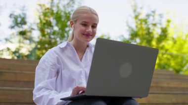 Genç bir kadın dışarıda dizüstü bilgisayar kullanarak oturuyor, gülümsüyor ve kablosuz kulaklıklarla kablosuz olarak dinleniyor..