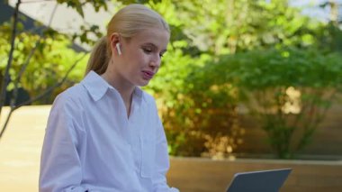 Kablosuz kulaklıklı, kendine güveni tam genç bir kadın. Laptopuyla video görüşmesi yapıyor. Açık havada, rahat bir ortamda oturuyor..
