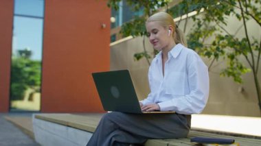 Modern bir ofis binasının yanında otururken bilgisayar ve kablosuz kulaklık kullanan profesyonel genç bir kadın..