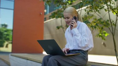 Gülümseyen genç bir kadın açık havada birden fazla iş yapıyor, telefonla konuşuyor ve modern bir şehir avlusunda dizüstü bilgisayar kullanıyor..