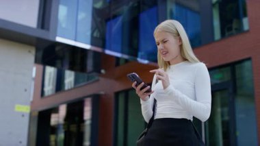 Şok olmuş genç bir kadın modern bir binanın önünde duruyor, akıllı telefonuna gözle görülür bir endişe ve acıyla bakıyor..