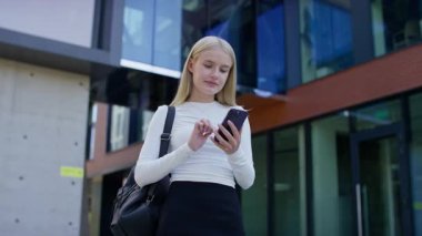 Modern bir binanın önünde duran genç bir kadın telefonuna hoşnutsuzluğun ve memnuniyetsizliğin ince bir ifadesiyle bakıyor..