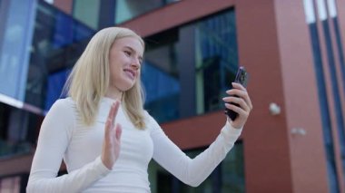 Modern bir binanın önünde gülümseyen bir kadın, akıllı telefon ekranına el sallayarak neşeli bir video görüşmesini bitiriyor..