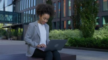İş kıyafetleri içinde gülümseyen bir kadın dışarıda modern binalarla otururken dizüstü bilgisayarı üzerinde çalışıyor..