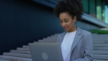 Gri ceketli profesyonel kadın, modern kentsel iş ortamında somut basamakların yanında dururken dizüstü bilgisayarına odaklandı..