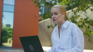 Kulaklık takan odaklanmış bir kadın, ağaçlarla dolu bir şehir ortamında dizüstü bilgisayarı üzerinde çalışırken bir video konuşması yapıyor..