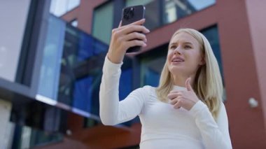 Kendine güvenen sarışın kadın, video selfie 'yi telefonla kaydediyor. Modern şehir binasının önünde baş parmağıyla işaret ediyor..
