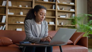 Spor ceketli genç profesyonel kadın, kitaplık ve bitkilerle süslü şık bir ofis salonunda dizüstü bilgisayara odaklanmış.