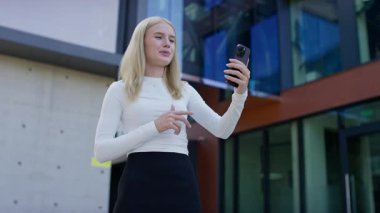 Modern bir binanın dışındaki video görüşmesi için akıllı telefon kullanan gülümseyen kız öğrenci çevrimiçi eğitimi, iletişimi ve dijital kampüs hayatını temsil ediyor..