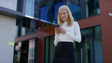 Modern bir binanın önünde durmuş gülümseyen genç bir kadın akıllı telefonuyla mesajlaşırken cep telefonu iletişimini, öğrenci hayatını ve dijital yaşam tarzını temsil ediyor..