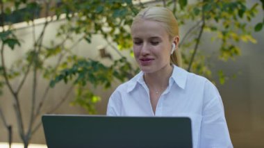 Modern bir ofis avlusunda çalışırken online toplantı için dizüstü bilgisayar ve kablosuz kulaklık kullanan mutlu bir iş kadını..