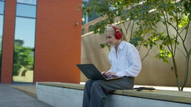 Modern bir üniversite binasının önünde oturmuş kulaklıkla müzik dinleyen genç bir kız öğrenci..