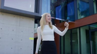 Modern üniversite binasının önünde duran genç kız öğrenci, video görüşmesi sırasında telefonla konuşurken gülümsüyor..