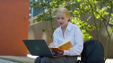 Dışarıda dizüstü bilgisayar ve not defteriyle oturan genç bir kız öğrenci modern kampüs ortamında online bir ders sırasında notlar alıyor..