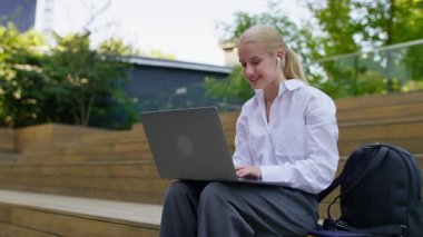 Ağaç merdivenlerde oturan gülümseyen genç bir kız öğrenci, modern kampüs ortamında kulaklıklı dizüstü bilgisayar kullanıyor..