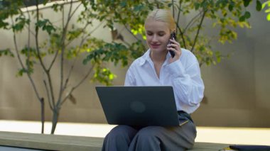 Akıllı telefonla konuşurken dizüstü bilgisayar kullanan gülümseyen genç iş kadını, modern bir şehir ortamında birden fazla iş yapıyor ve uzaktan çalışıyor..