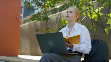 Çevrimiçi derslere katılan, dizüstü bilgisayar kullanırken elinde not defteri ve kalem tutan, modern şehir ortamında uzaktan eğitim gören odaklanmış genç bir kadın..