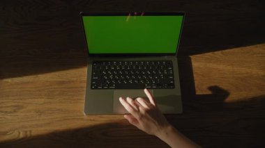 Sıcak güneş ışığı altında, ahşap masadaki yeşil renkli anahtar ekranlı dizüstü bilgisayarda kayan bir elin üst görüntüsü.