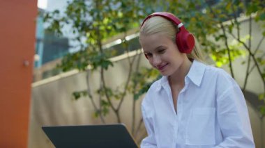 Kırmızı kulaklık takıp dışarıda dizüstü bilgisayar kullanan gülümseyen genç bir kadın uzaktan çalışma, internet iletişimi, müzik ve modern dijital yaşam tarzını gösteriyor..