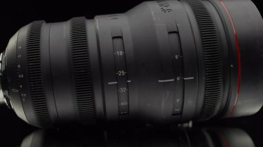 İşaretli odaklanma ve yakınlaştırma ölçekleri ile profesyonel bir yakınlaştırma lensinin yan görüntüsü, koyu yansıtıcı bir yüzey üzerinde görüntülenir.