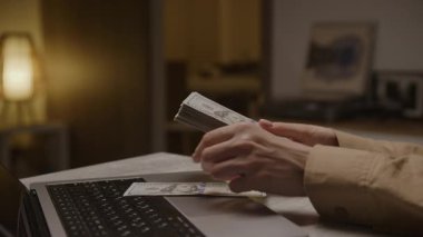 El ele tutuşup, rahat bir iç mekanda, kişisel finans, çevrimiçi gelir ve bütçeyi gösteren bir dizüstü bilgisayarın yanındaki Amerikan doları banknotlarını saymak..