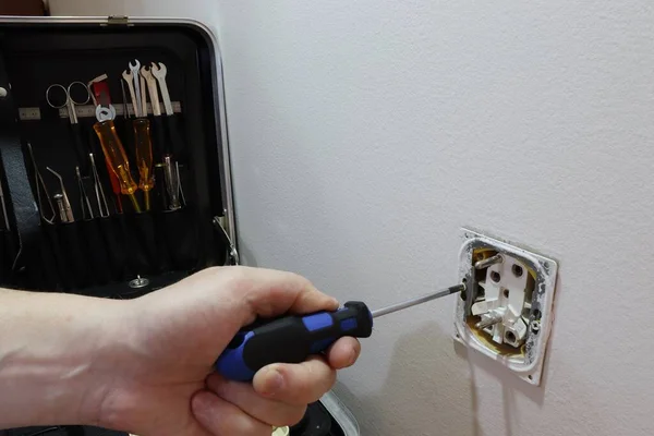 An electrician repairs an old electrical outlet. The electrician removes the electrical outlet. In the background a box with tools.