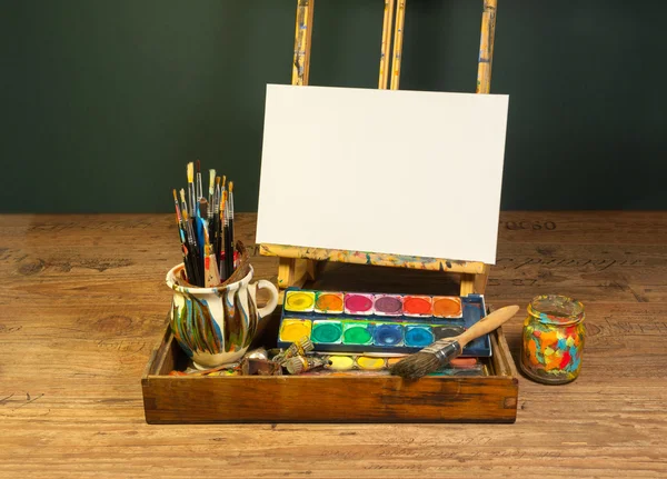 Paleta de caballete acuarelas y pinceles con lienzo blanco vacío en el estudio de arte pintor — Foto de Stock