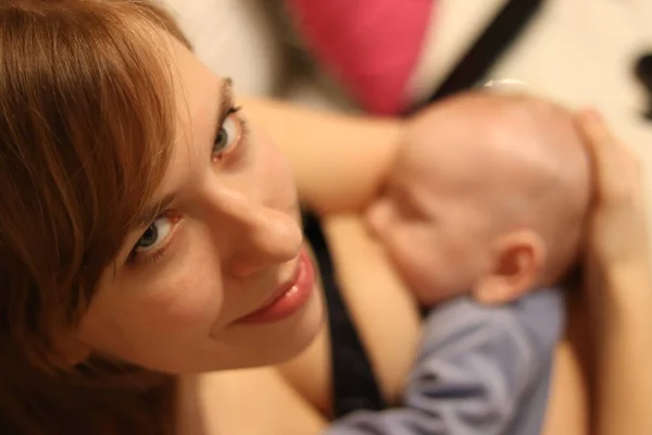 Beyaz kadın çocuğunu emziriyor ve kameraya gülümsüyor.