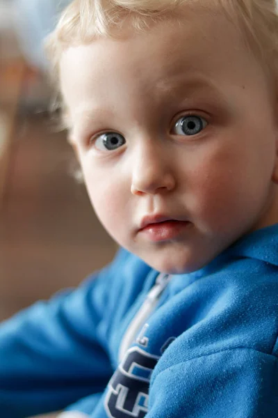 Mavi gözlü, sarı, kıvırcık saçlı, mavi kapüşonlu ve kameraya bakan çocuk.